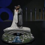 Two intricately dressed fantasy figurines standing closely on a rocky pedestal, a man and woman facing each other as if about to kiss, with a carved waterfall beneath them.