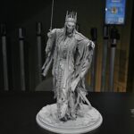 Grey statue of a regal crowned figure in tattered robes, holding a sword, standing on a cracked pedestal.