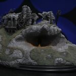 Miniature rocky hill with ruined stone arches atop a diorama pedestal against a dark blue background.