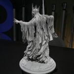 Gray statue of a crowned, flowing-robed figure reaching forward, with ribbon-like strands, mounted on a round base.