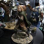 Battle-ready fantasy figurine: young woman in green armor and cloak, blonde hair, sword drawn on a rocky diorama base