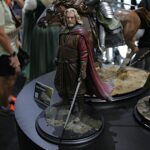 Fantasy action figures on circular black display bases at a convention, featuring a bearded wizard in red cape centerpiece on a rocky diorama.
