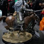 Armored knight figure riding a horse on a display pedestal at a convention, detailed metal armor with a long sword; crowd in the background observing the exhibit