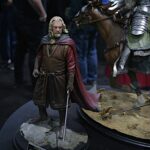 Figure of a medieval-styled man in brown robes with a cape and sword, standing on a rocky display, next to a armored horse figure on a circular pedestal at a convention show