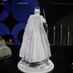 Back view of a gray Batman action figure wearing a cape, standing on a rocky pedestal on a display shelf with a dark background and blue circular shapes behind it.