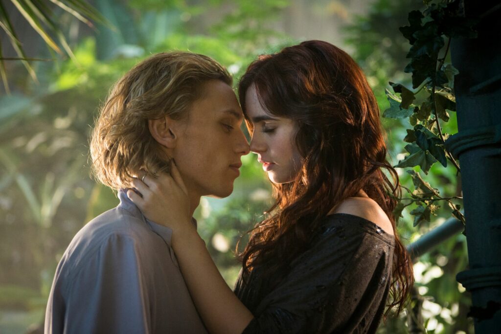 Two women standing close, about to kiss, in a sunlit garden with greenery.