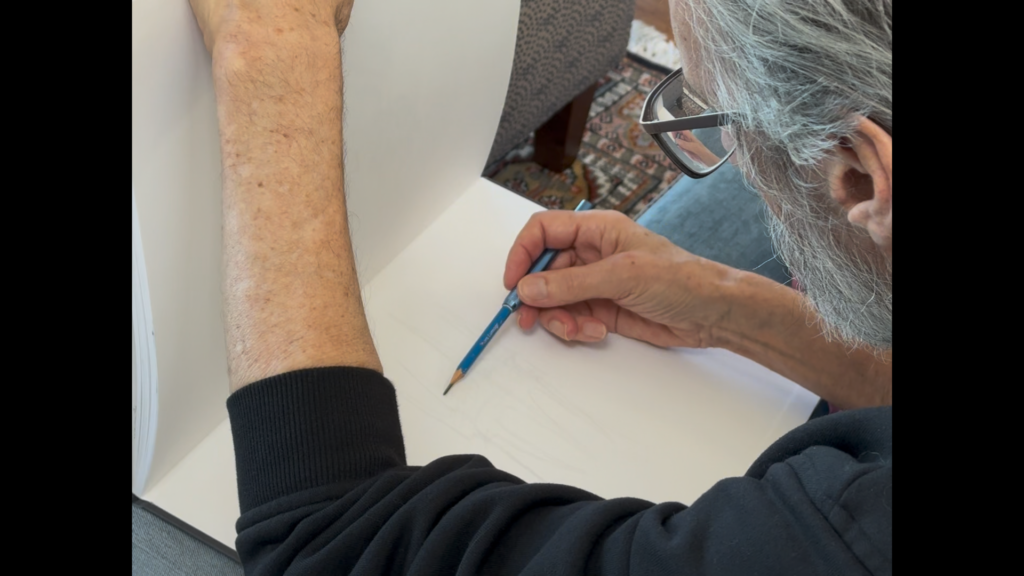 Close-up of an elderly person wearing glasses drawing with a blue pencil on white paper, viewed from above.