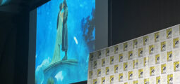 Large stage screen shows two people kissing on a blue backdrop, with Comic-Con panels along the foreground wall nearby.