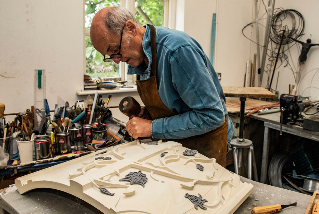 Artist Stephen Broadbent works on a prototype carving of part of Tolkien's memorial. He is working with a hammer and chisel, on an intricate design of weaving vines and flowers. 