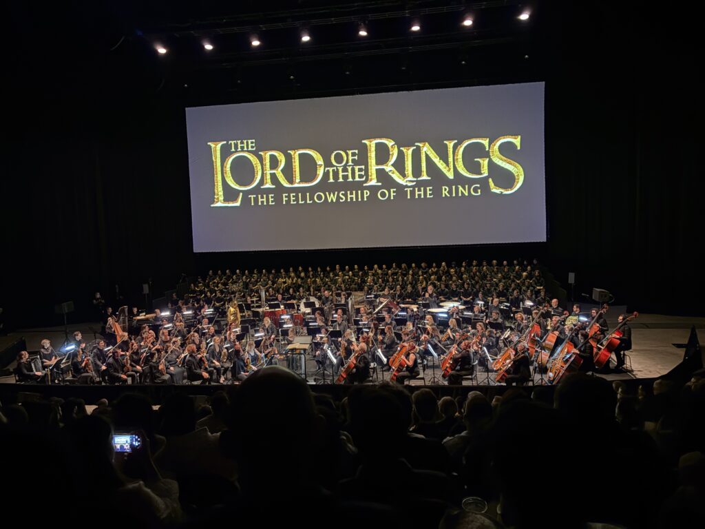 The full orchestra, with choir assembled behind, are ready to play in Sydney. On a screen behind and above them is the title care for The Lord of the Rings: The Fellowship of the Ring.