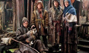 Man in a hooded cloak sits with a cat, while three children and a woman in fur robes stand beside him in a rustic wooden village setting.