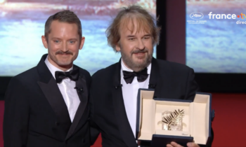 Two men in tuxedos smile on stage, holding an open award case with a palm-leaf trophy.