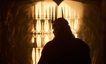 Silhouette of a person wearing a tall spiked crown against a glowing orange gate light in the background.