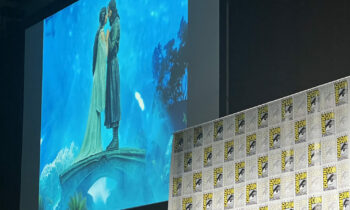 Large stage screen shows two people kissing on a blue backdrop, with Comic-Con panels along the foreground wall nearby.